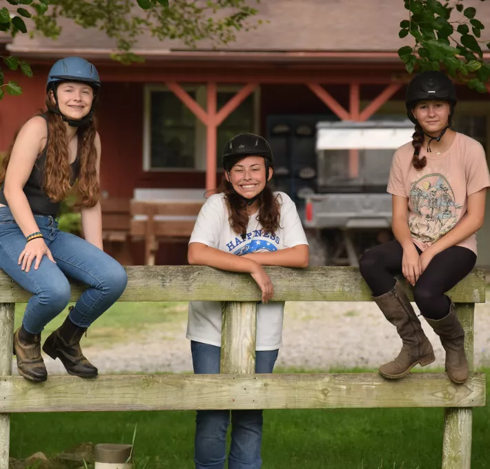 Two campers sitting on a fence with one standing between them. All wear riding helmets